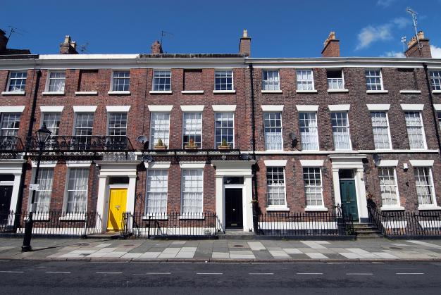 Row of classic Georgian-style townhouses with red brick facades and white-trimmed sash windows. The buildings are three stories tall with black wrought-iron railings along the front entrances. Each townhouse has a distinctively colored front door, including yellow, black, and dark green. The street in front is paved with a sidewalk, and a traditional black street lamp is visible on the left. The sky is clear and blue, casting bright sunlight on the buildings.