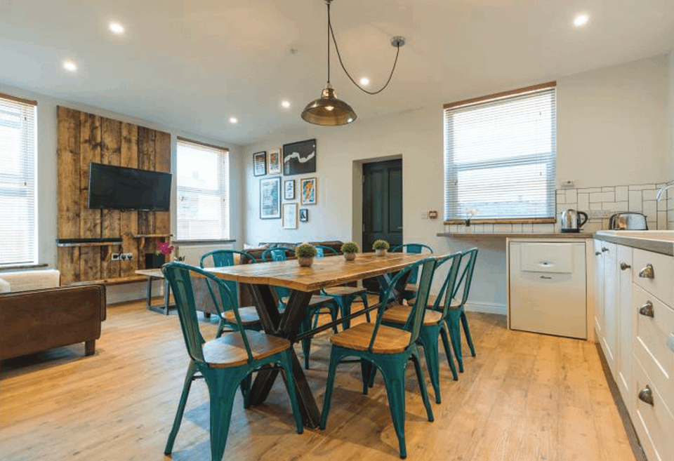 Bright, open-plan kitchen and dining area with a modern, rustic design. The space features a large wooden dining table surrounded by teal metal chairs with wooden seats. Light wood flooring complements white kitchen cabinets with silver handles, a countertop, and appliances including a kettle and toaster. A mounted TV and wood-paneled accent wall are on one side, while framed artwork decorates another wall. Natural light streams through multiple windows, creating a warm, inviting atmosphere.