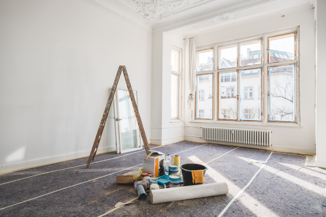 Bright, empty room undergoing renovation with large windows letting in natural light. The room has ornate ceiling moldings, white walls, and a radiator beneath the windows. A wooden A-frame ladder leans against the wall, and painting supplies—including paint cans, a roller, a bucket, and a roll of protective paper—are scattered on the floor, which is covered with protective material.