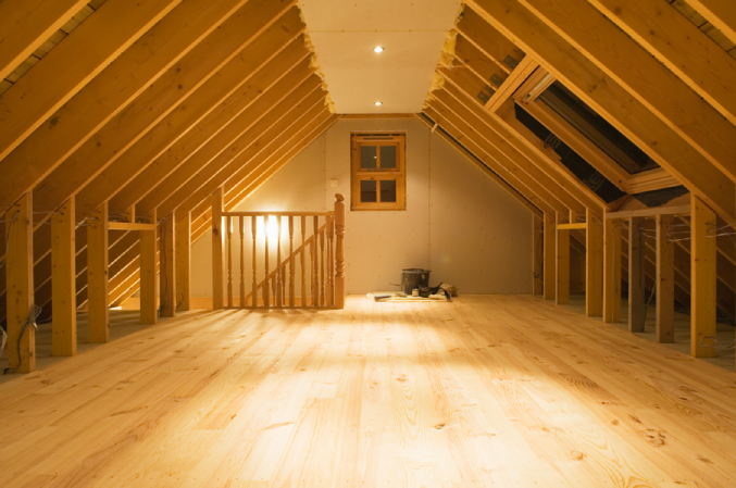 Interior view of a newly finished attic space with exposed wooden beams and a light wood floor. The attic has a sloped ceiling with recessed lighting, a small wooden-framed window at the far end, and a skylight on the right side. A wooden railing surrounds the staircase opening, and some construction materials are placed near the back wall. The space is clean, well-lit, and appears ready for use or further furnishing.