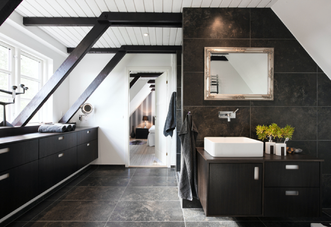 Modern attic bathroom with a sleek, minimalist design featuring dark tile flooring and walls contrasted by white ceilings and fixtures. Exposed dark wooden beams run along the sloped ceiling. The bathroom includes a rectangular vessel sink on a dark wood vanity, a large mirror above it, and neatly folded towels on a long countertop by the windows. Natural light streams through the large windows, highlighting the clean, contemporary aesthetic, with a glimpse of a bedroom visible through an open doorway.