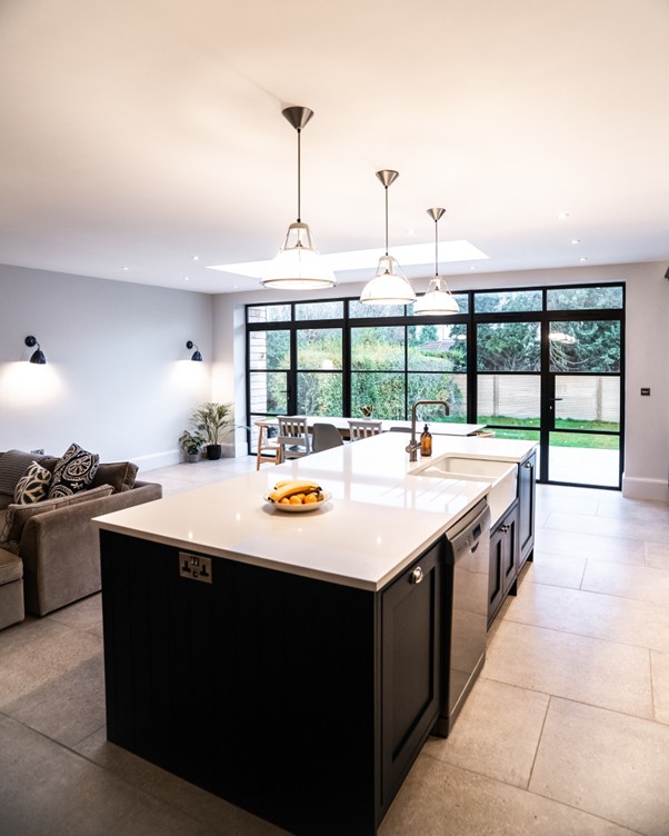 Modern open-plan kitchen and living area with a large white island, black cabinetry, pendant lights, gray tiled floors, and floor-to-ceiling glass doors opening to a garden patio.