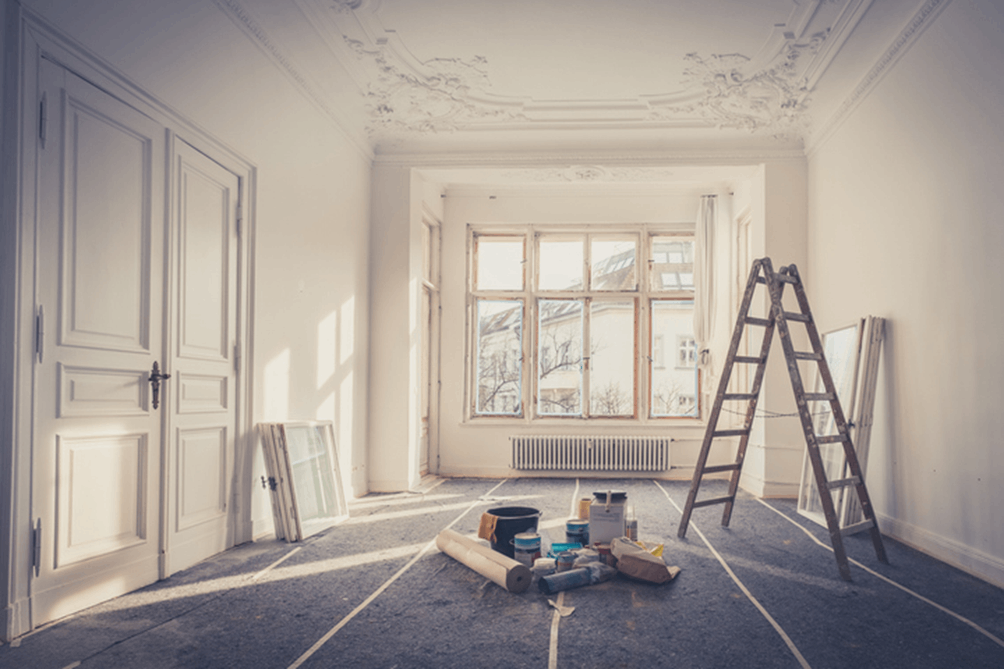 Sunlit room undergoing renovation with white walls and ornate ceiling moldings. The floor is covered with protective material, and painting supplies—including paint cans, a roller, a bucket, and a roll of paper—are scattered in the center. A wooden A-frame ladder leans against the wall, and several window frames rest nearby. Large windows at the far end let in natural light, highlighting the room’s high ceilings and decorative details.