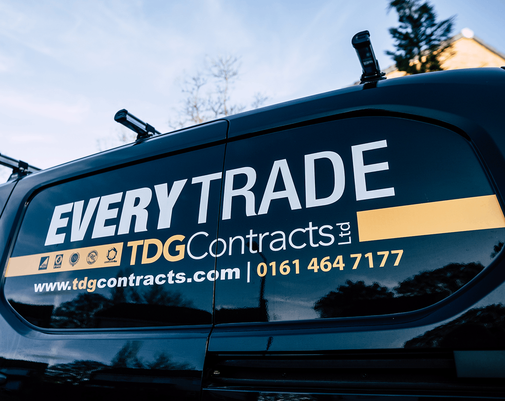 Close-up view of the side of a dark-colored company van displaying branding for “EVERYTRADE TDG Contracts Ltd.” The text includes the company website, www.tdgcontracts.com , and a contact phone number, 0161 464 7177. Several small certification or service icons are shown above the company name, and the van roof is equipped with roof racks. The vehicle reflects the surrounding trees and sky.