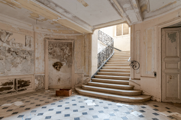 Abandoned interior of a grand building with peeling paint and damaged walls, featuring a tiled floor scattered with debris and a curved staircase with an ornate railing leading up toward a bright upper level.