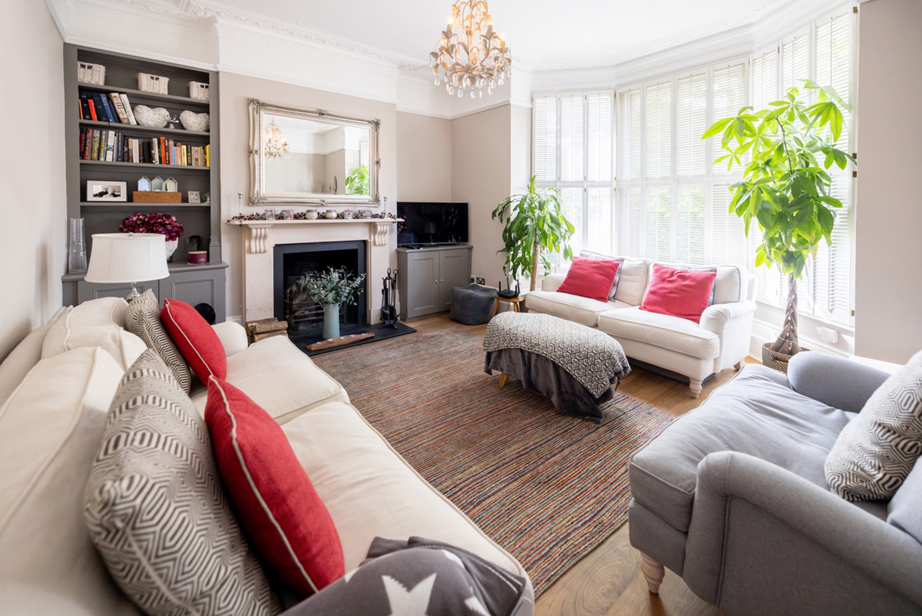 Bright, spacious living room with cream sofas, a grey armchair, striped rug, decorative fireplace with mirror above, built-in shelving, large bay windows with white blinds, and indoor plants.