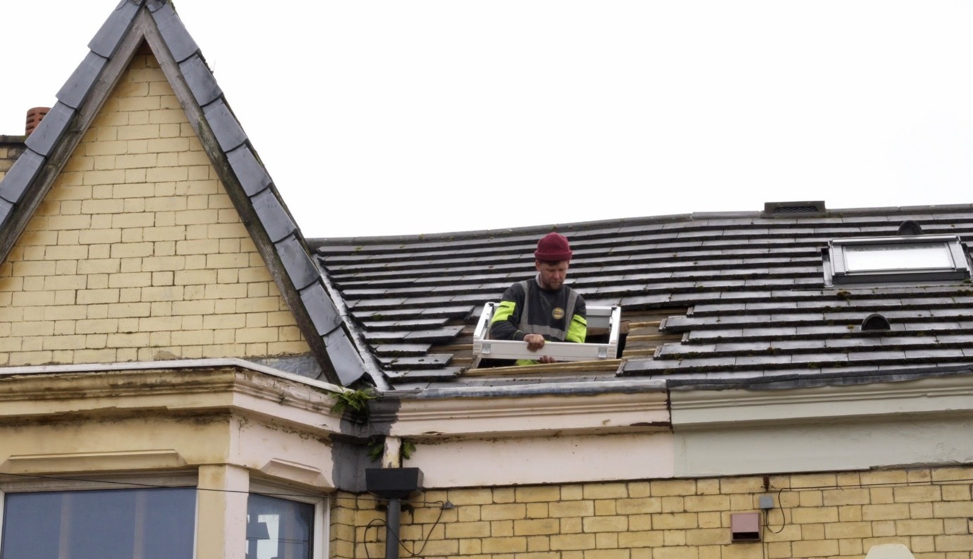 A construction worker wearing a red beanie and high-visibility clothing installs or adjusts a roof window while standing on the tiled roof of a brick house under an overcast sky.