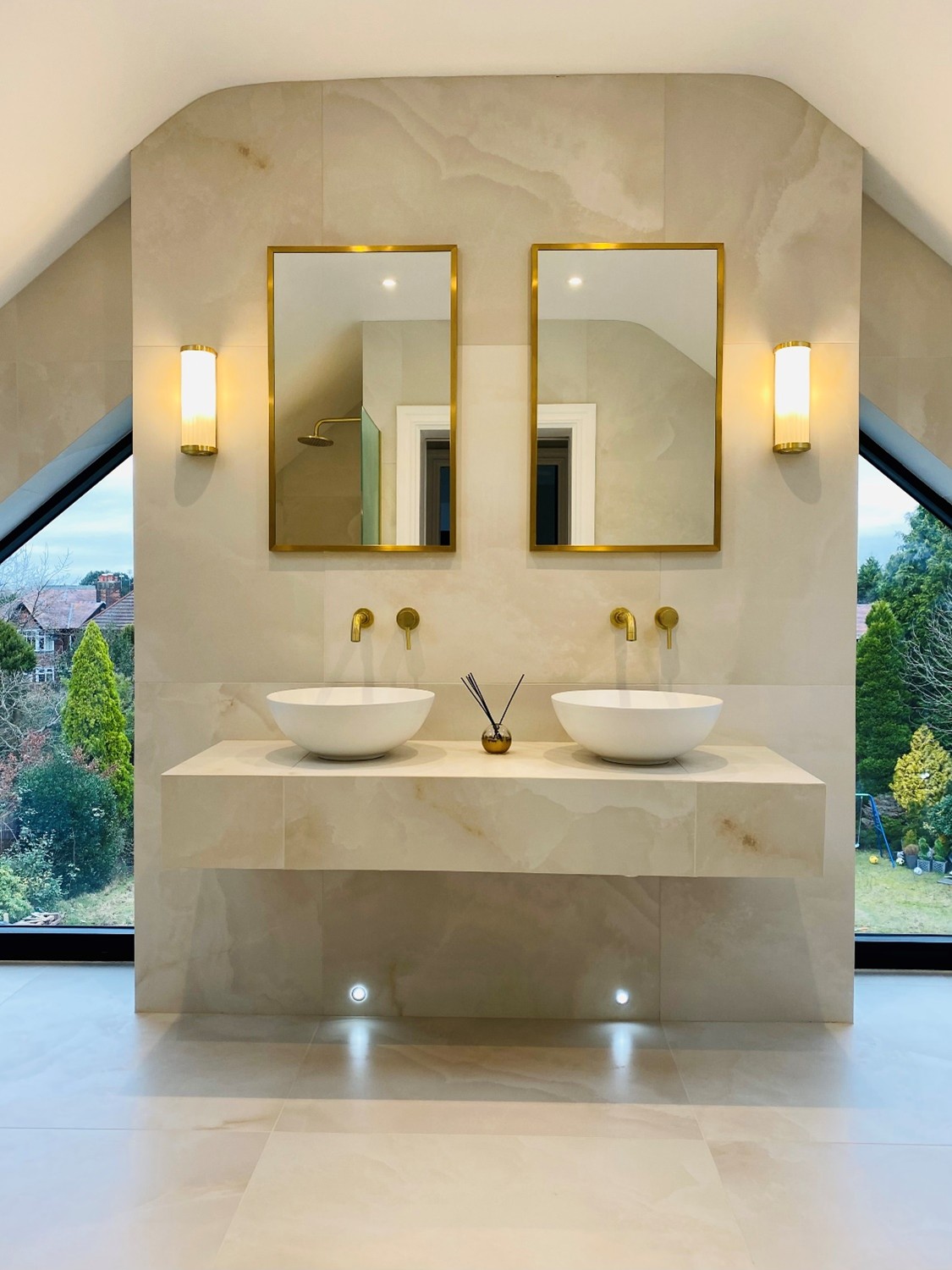 A modern bathroom with two white vessel sinks mounted on a floating stone vanity, paired with gold wall-mounted taps and matching framed mirrors. Soft wall lights and large angled windows on either side create a bright, minimalist space with views of trees outside.