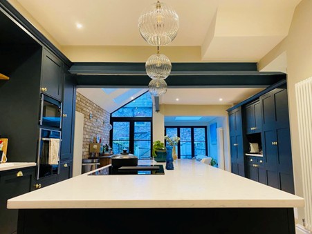 A modern kitchen with dark navy cabinets, a large white island countertop in the foreground, and three glass pendant lights hanging overhead. The space features exposed brick on one wall, recessed ceiling lighting, and large black-framed windows and doors at the back letting in natural light.