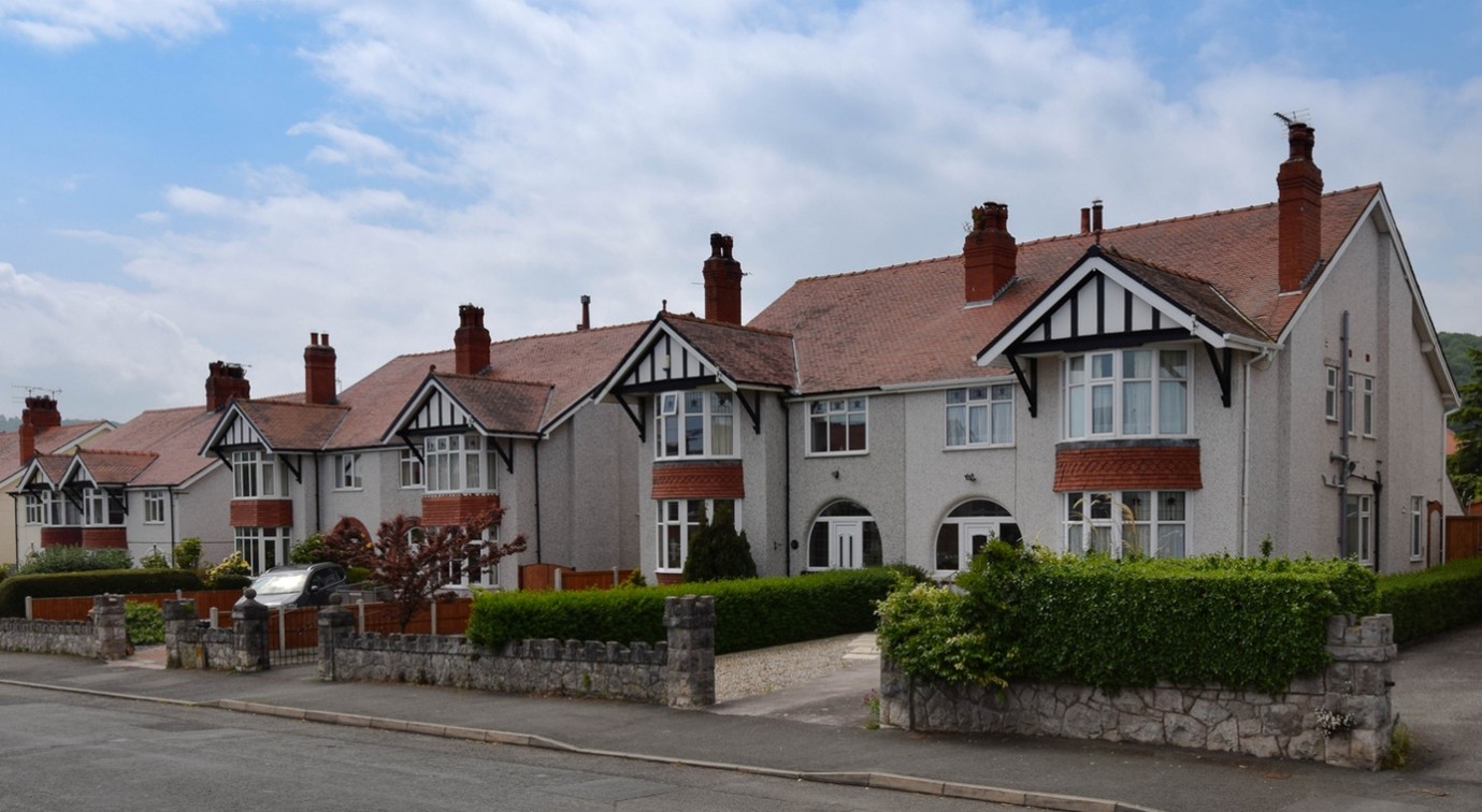 A row of semi-detached suburban houses with white pebble-dash walls, red tiled roofs, black timber trim, and neatly trimmed hedges along a quiet residential street under a cloudy sky.