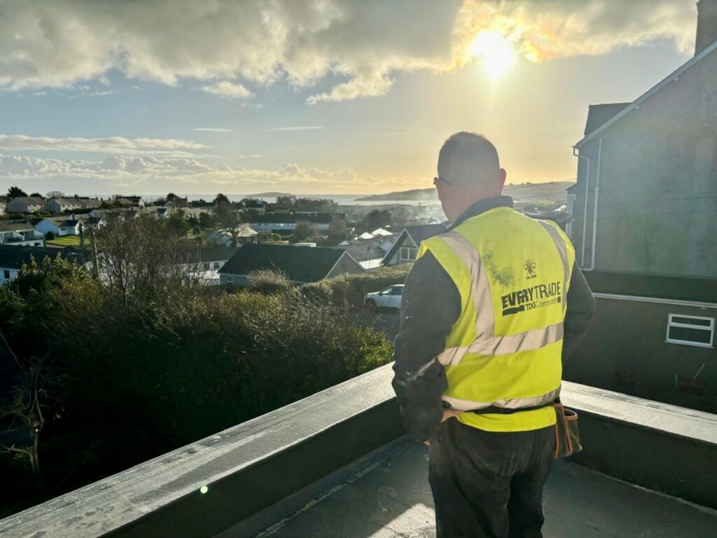 Construction worker in a high-visibility vest standing on a balcony or flat roof, overlooking a residential neighborhood and distant coastline under a bright, cloudy sky.