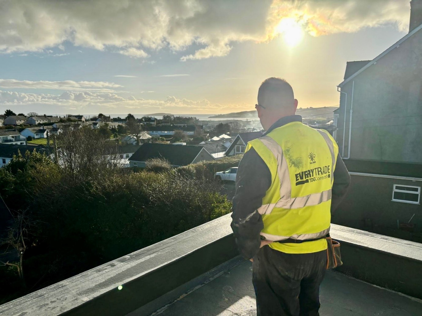 Construction worker in a high-visibility vest standing on a balcony or flat roof, overlooking a residential neighborhood and distant coastline under a bright, cloudy sky.