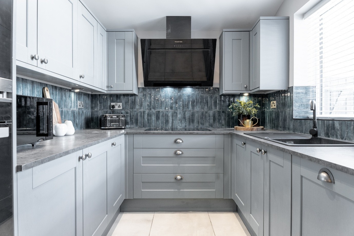 Contemporary U-shaped kitchen with light gray shaker cabinets, gray countertops, a glossy blue-gray tiled backsplash, black extractor hood, integrated sink beneath a window with white blinds, and light tiled flooring.