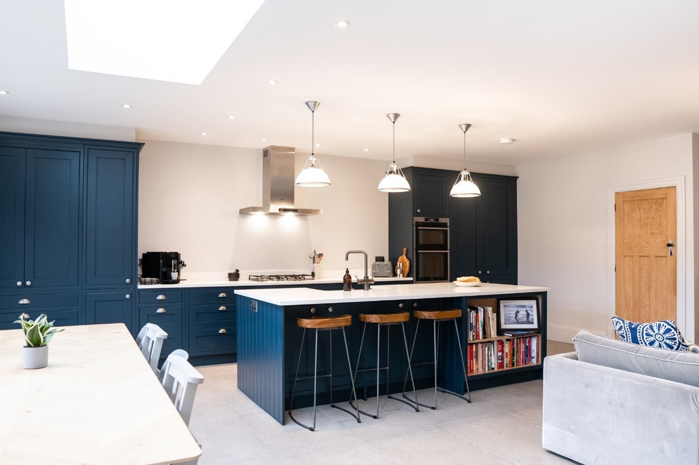 Contemporary open-plan kitchen featuring navy blue shaker-style cabinets, a white island with breakfast seating and built-in shelving, pendant lighting, a skylight, integrated appliances, and an adjoining dining and living area