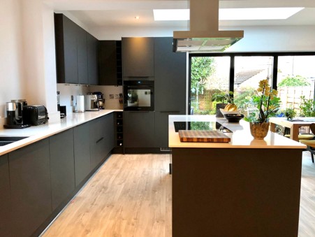 "Modern kitchen with matte dark-gray cabinets, white countertops, a central island with a built-in cooktop and stainless steel range hood, light wood flooring, and large sliding glass doors opening to a garden