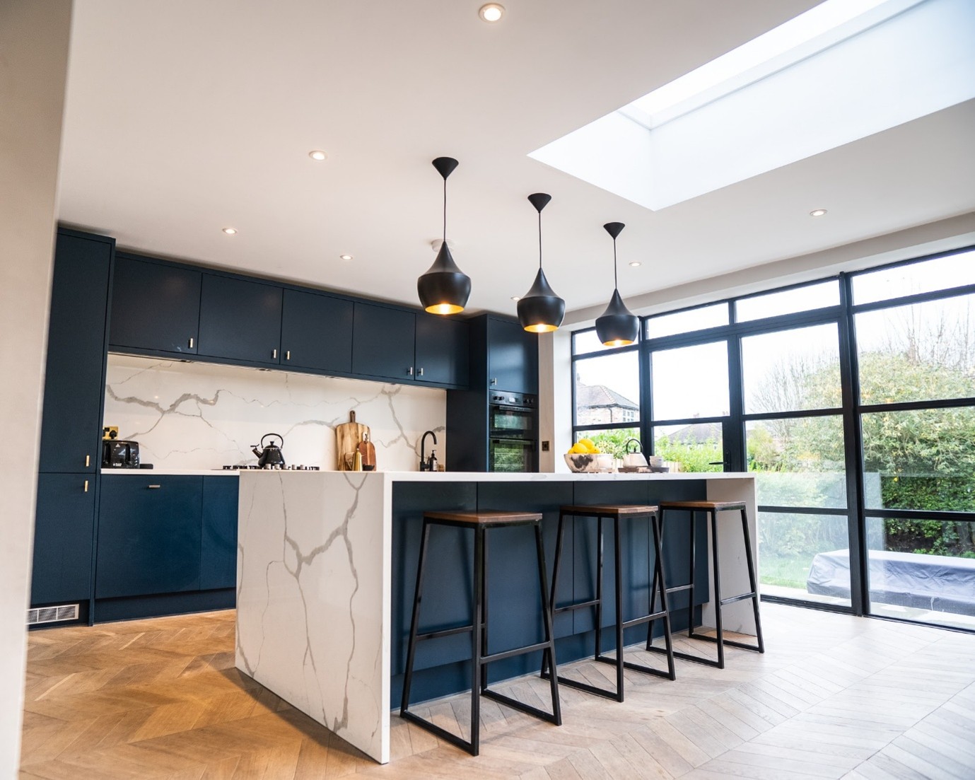Modern open-plan kitchen with navy blue cabinets, a white marble waterfall island with three bar stools, black pendant lights, a large skylight, and floor-to-ceiling black-framed windows overlooking a garden