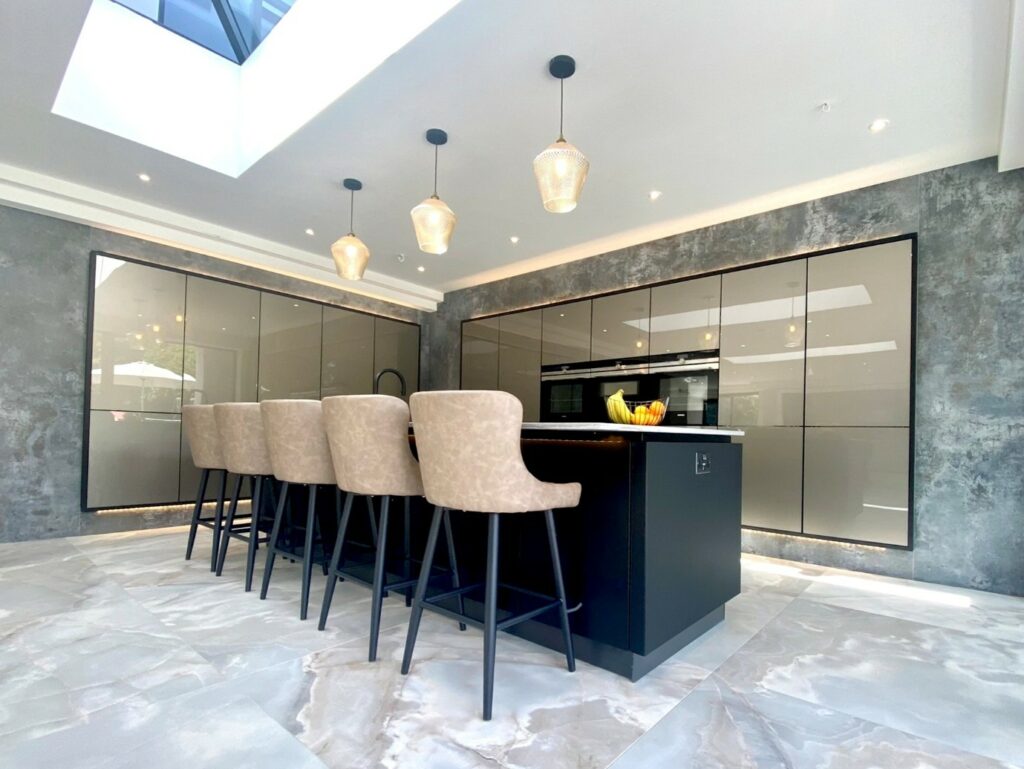 Spacious contemporary kitchen with glossy handleless cabinets, a large black island with upholstered bar stools, pendant lighting, marble-effect floor tiles, and a skylight providing abundant natural light
