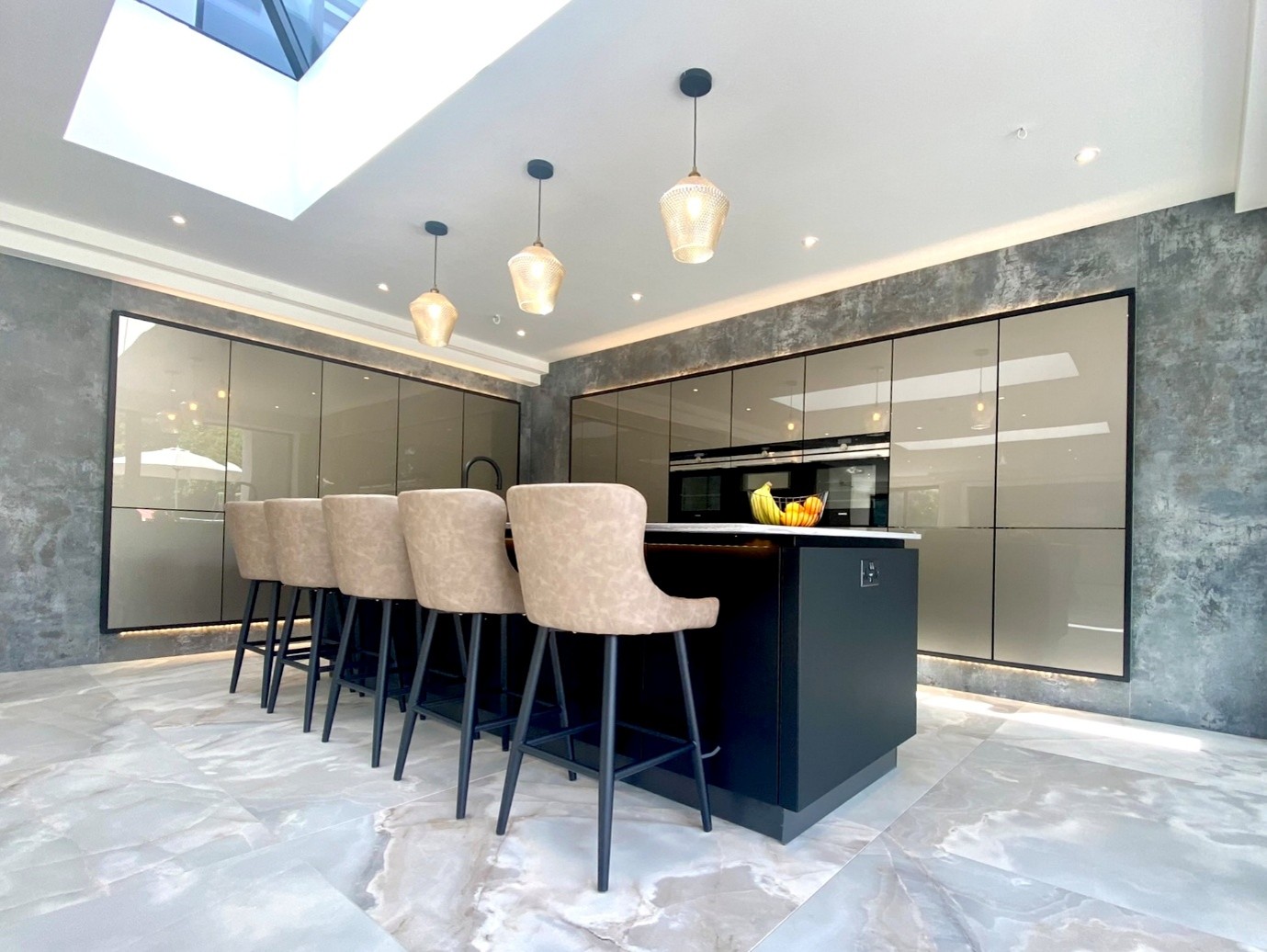 Spacious contemporary kitchen with glossy handleless cabinets, a large black island with upholstered bar stools, pendant lighting, marble-effect floor tiles, and a skylight providing abundant natural light
