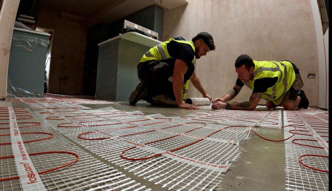 Two workers wearing high-visibility vests kneel on a concrete floor, installing electric underfloor heating mats with red heating cables in a partially finished room. Cabinets and construction materials are visible in the background.
