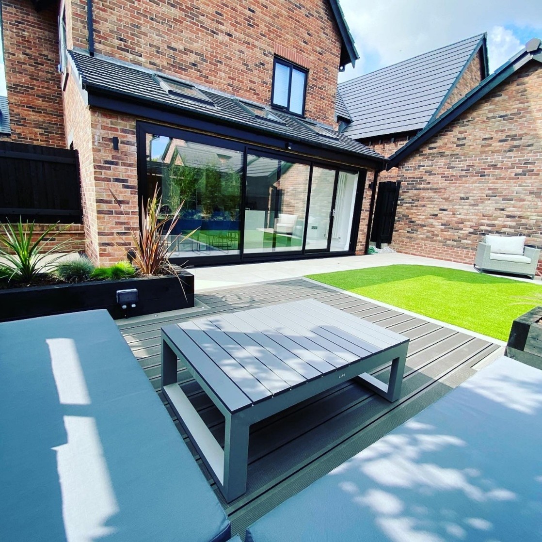 Contemporary brick house extension with large black-framed sliding glass doors opening onto a modern landscaped garden. The outdoor space features grey composite decking, raised black planters, minimalist patio furniture, and a bright green lawn, creating a clean and stylish entertaining area in daylight.