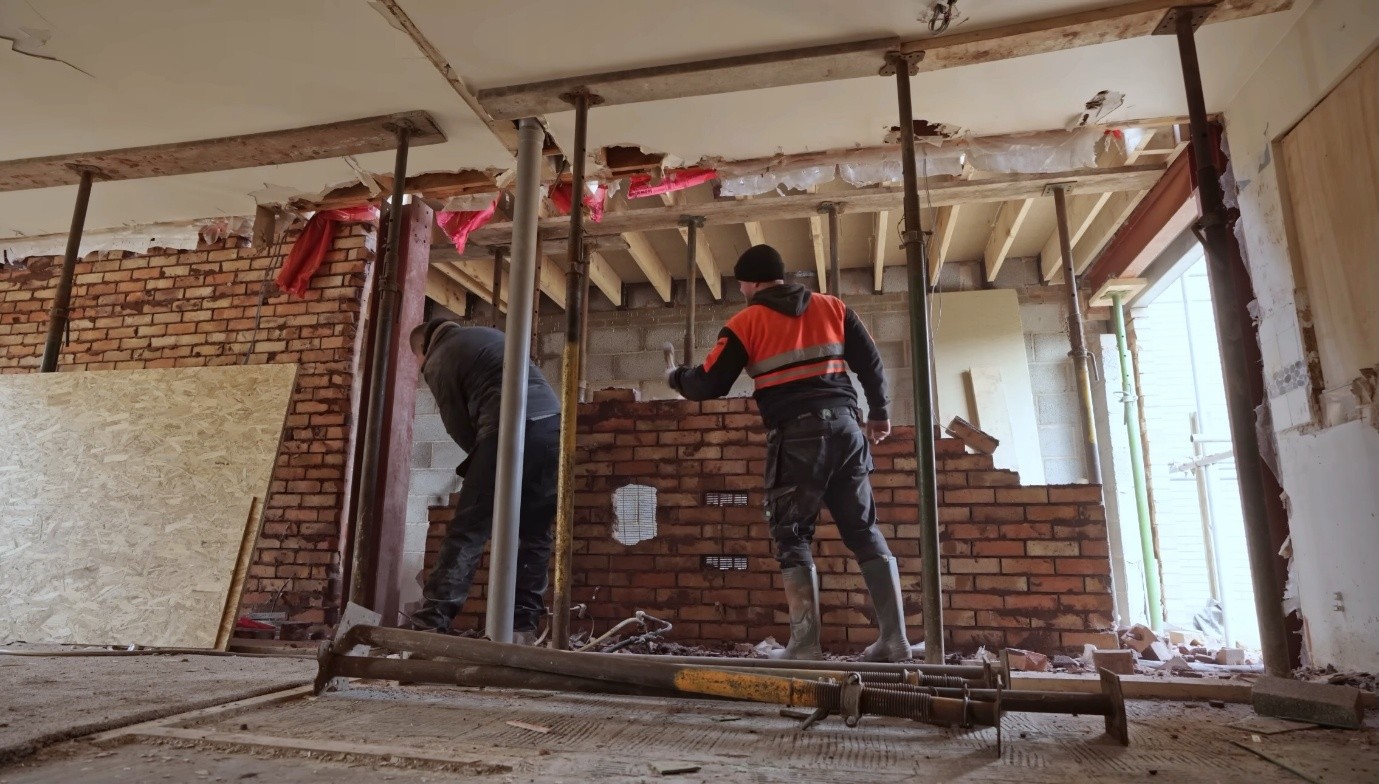 Interior structural renovation in progress, showing builders removing an internal brick wall beneath exposed ceiling joists. Temporary steel support props and beams are installed to support the structure, with construction debris on the floor and partially demolished walls visible throughout the open-plan space.