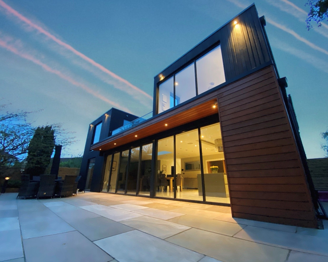 Modern two-storey contemporary home at dusk with dark timber and metal cladding, large floor-to-ceiling glass doors, a first-floor balcony, and warm interior lighting opening onto a spacious light stone patio. The house is photographed from a low angle against a blue evening sky streaked with pink clouds.