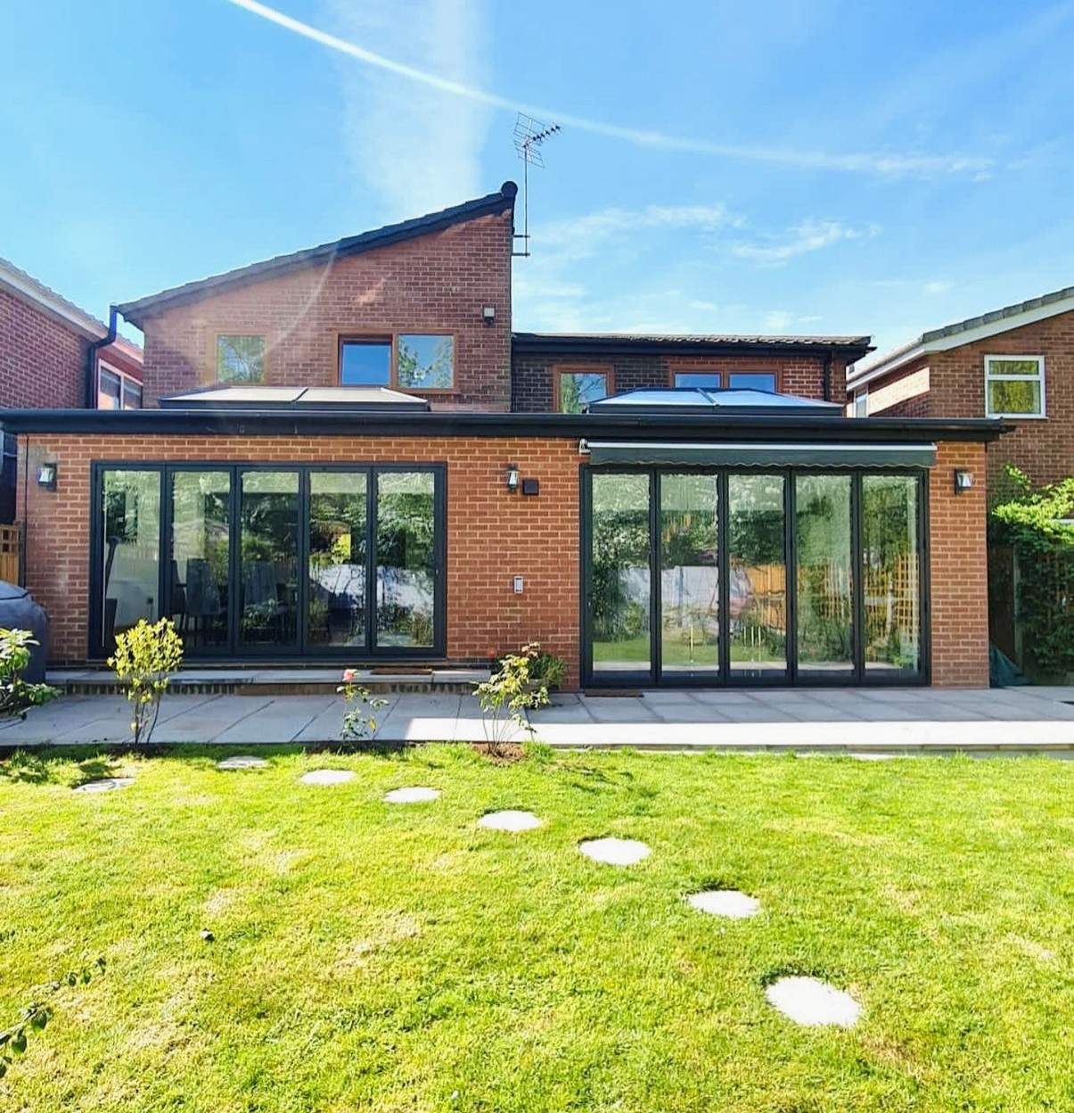 Rear view of a detached brick home featuring a modern single-storey extension with black-framed bi-fold doors and large roof lanterns. The extension opens onto a spacious patio and well-maintained lawn with stepping stones, creating a bright and contemporary indoor-outdoor living space on a sunny day.