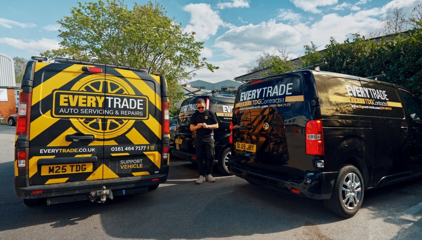 Three branded Everytrade service vans parked in a company yard on a sunny day. The vehicles display black and yellow livery promoting auto servicing, repairs, and trade services, while a staff member stands between the vans. Trees, industrial buildings, and a blue sky with scattered clouds form the background.