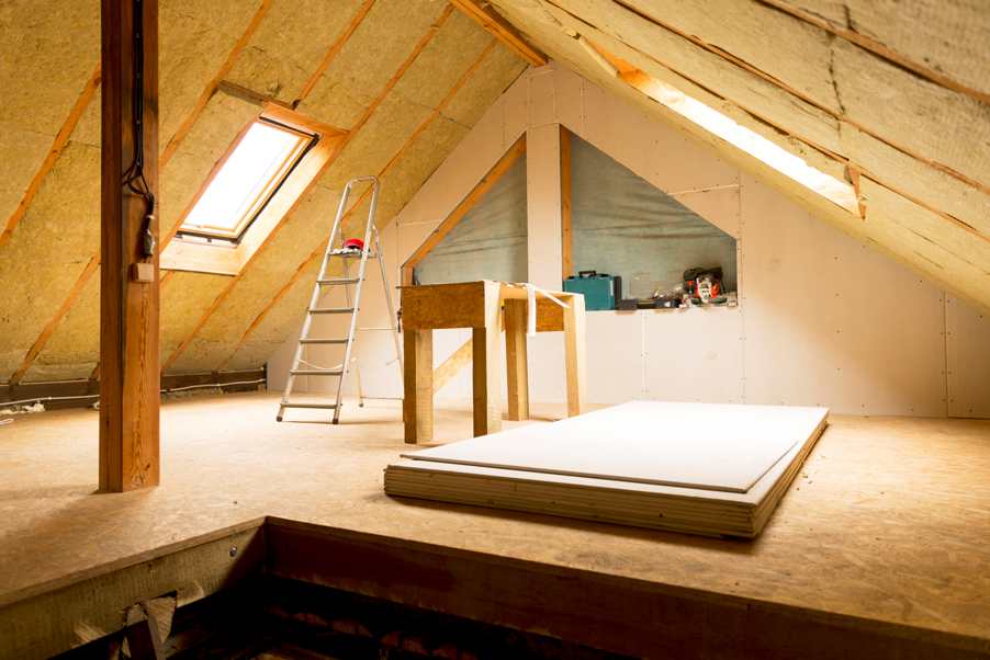 Attic conversion under construction with sloped insulated roof panels, a roof window providing natural light, exposed timber supports, and newly installed plasterboard walls. A step ladder, workbench, tools, and stacked plasterboards are visible on the unfinished timber floor, showing the loft space in the process of being transformed into a habitable room.