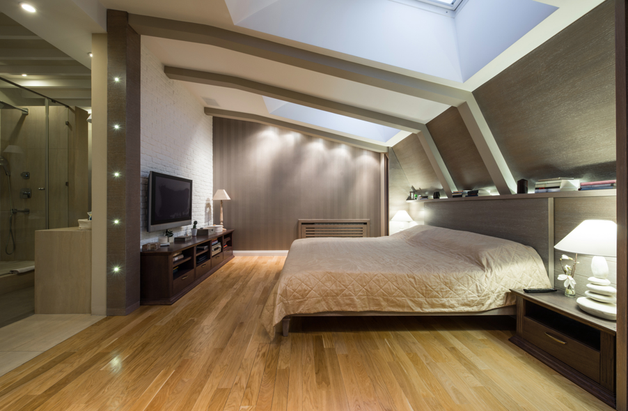 Modern attic-style bedroom with sloped ceilings and skylights, featuring a large bed with beige bedding, warm wood flooring, bedside lamps, a wall-mounted TV on a dark wood media unit, and an open ensuite bathroom with a glass shower visible to the left.