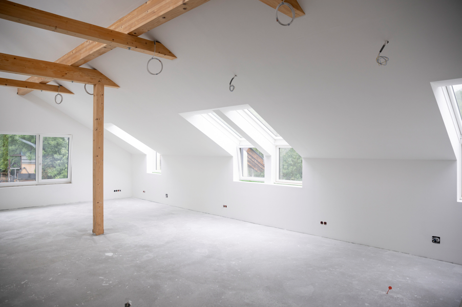 Unfinished loft room with white plastered walls, exposed timber beams and support posts, sloped ceilings with multiple skylights, hanging electrical cables for future lighting, and large windows providing natural light. The room has a bare concrete floor and is ready for final fit-out and decoration.