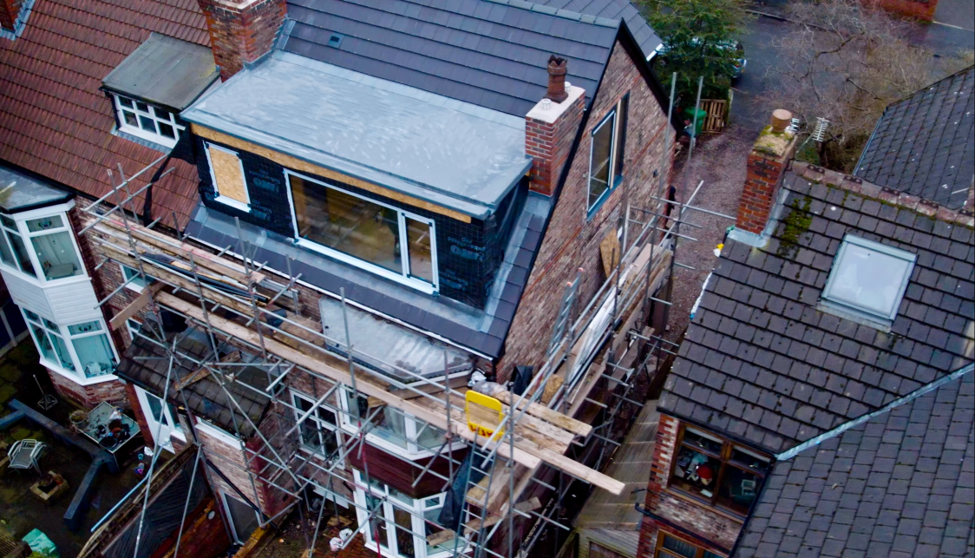 Aerial view of a house undergoing a loft conversion and roof extension, with scaffolding surrounding the property. Newly installed dormer windows and roofing materials are visible, while neighbouring residential buildings closely border the construction site.