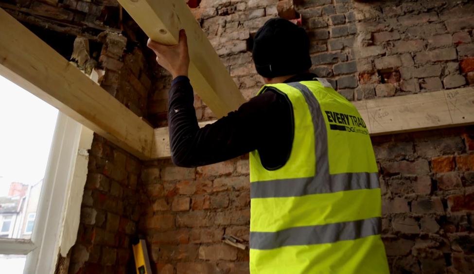 Construction worker in a high-visibility vest installing a structural timber beam inside a property renovation. Exposed brick walls, timber supports and an unfinished interior highlight ongoing building work during a loft conversion or home extension project.