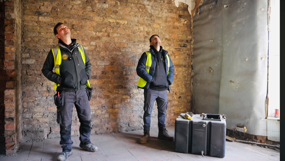 Two construction workers stand inside a stripped-back property during a building renovation. Exposed brick walls, unfinished flooring, insulation materials and site equipment are visible as the workers inspect the room before further construction work.