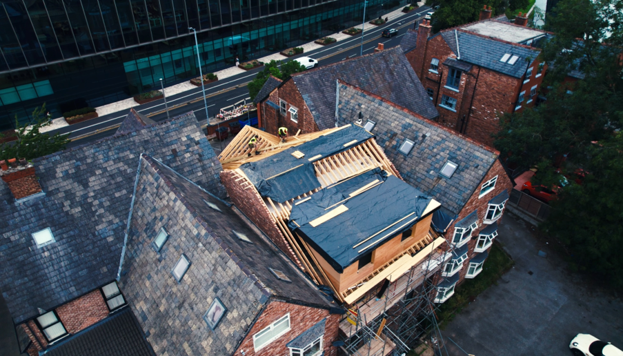 Aerial view of a large brick residential building undergoing a roof extension, with exposed timber framing, waterproof sheeting, and scaffolding surrounding the construction area. Adjacent slate roofs and a modern glass-fronted office building are visible in the background, with roads and parked vehicles nearby.