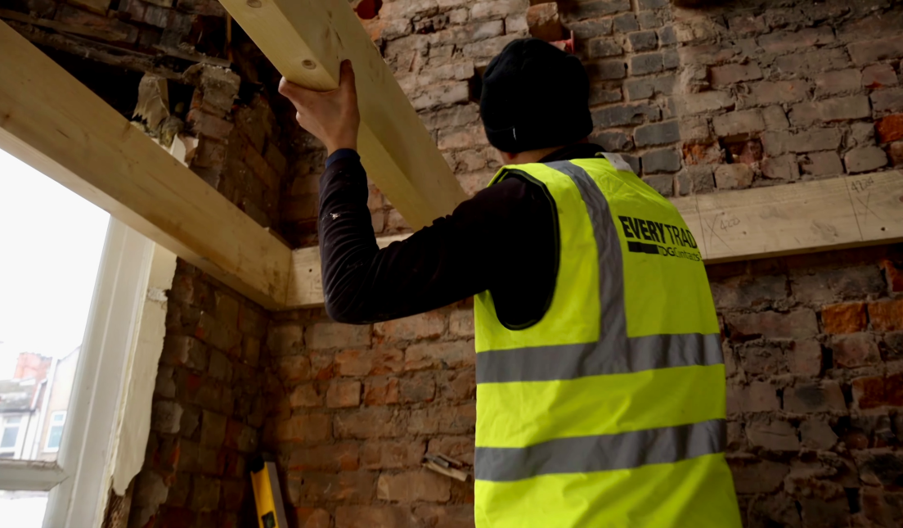 Construction worker wearing a high-visibility vest positions a timber beam within a partially renovated room. Exposed brick walls, newly installed timber supports, and an open window reveal ongoing structural refurbishment work inside the building. The high-visibility vest displays the company branding "Everytrade Lofts & Extensions