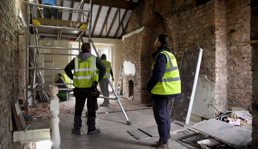 Interior renovation project in progress within a large brick building, featuring exposed brick walls, partial plasterwork, and scaffolding erected beneath an open roof structure. Several construction workers wearing high-visibility vests are inspecting and carrying out building works, while construction materials and debris are visible throughout the space