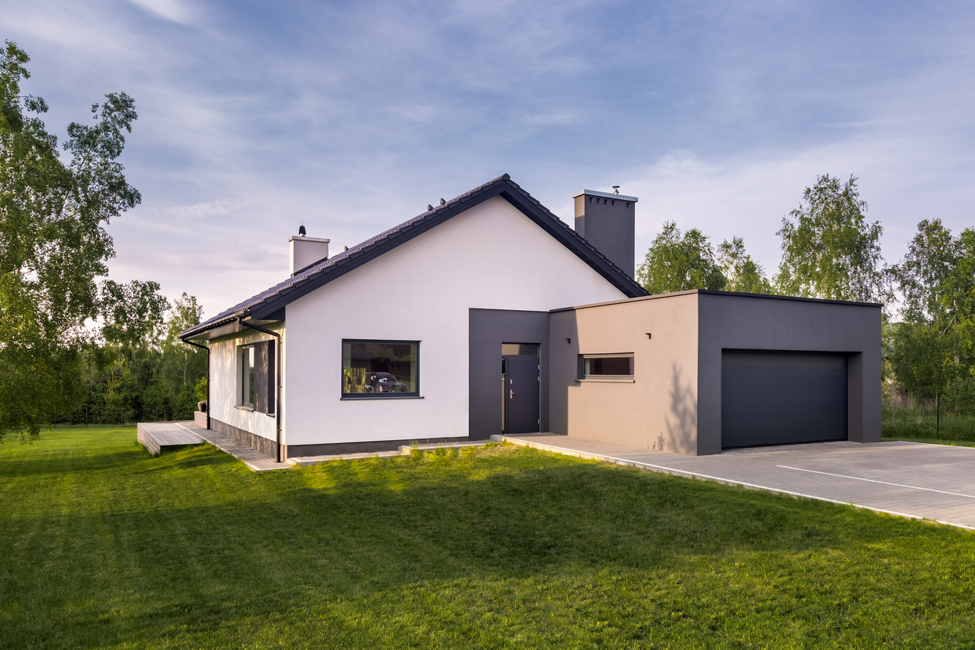 Modern detached family home with a white rendered exterior, dark grey garage, pitched roof, and landscaped lawn, set within a spacious plot surrounded by mature trees.