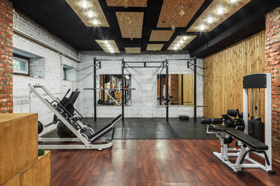 Modern gym and fitness studio featuring strength-training equipment, including a squat rack, leg press machine, weight bench, and free weights, with exposed brick walls, timber panelling, and rubber flooring.
