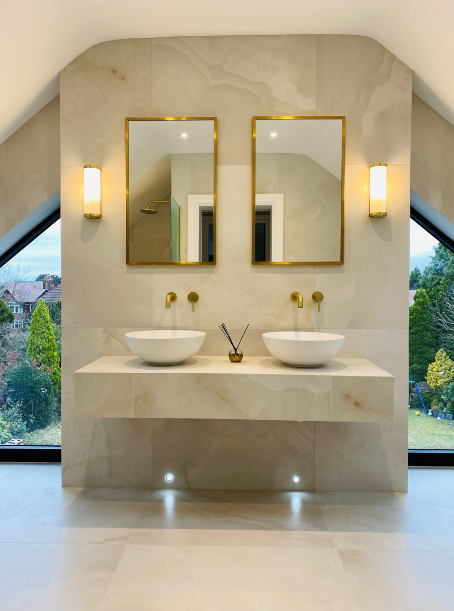 Modern luxury bathroom featuring a floating vanity with two white vessel sinks, wall-mounted brass taps, and matching brass-framed mirrors. Large light-stone tiles cover the walls and vanity, while wall sconces and recessed floor lights provide soft illumination. Full-height windows on either side offer views of a landscaped garden, and the vaulted ceiling enhances the spacious feel