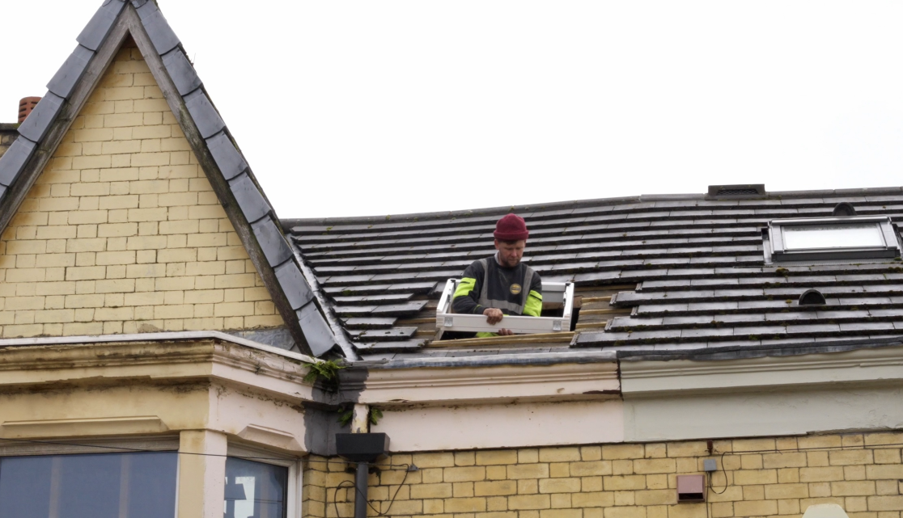 Person positioned at a roof opening while installing or adjusting a roof window on a tiled pitched roof. The yellow-brick building features multiple rooflights, guttering, and decorative gable detailing beneath an overcast sky