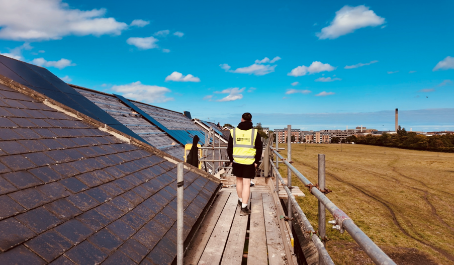 Person wearing a high-visibility vest walking along scaffolding beside a pitched slate roof during a roofing project. The roof includes sections under renovation and roof windows, with an open grassland area, residential buildings, and a blue sky with scattered clouds in the background